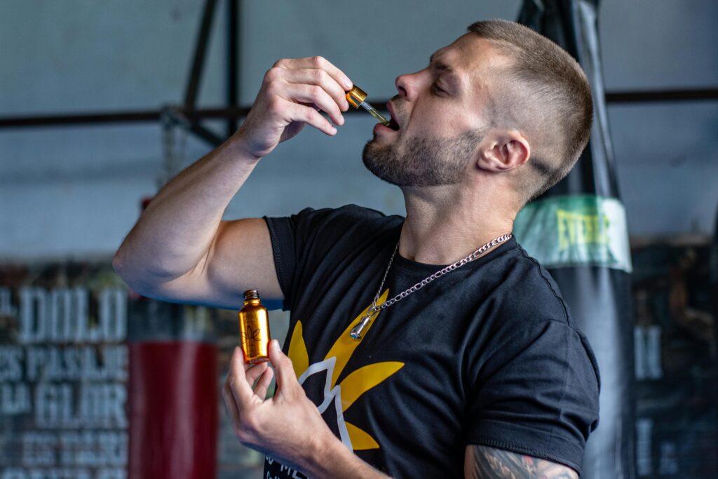 Adult man consuming hemp oil with a dropper in a gym. Emphasizes masculinity and wellness.